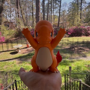 May include: Orange Charmander figurine with outstretched arms and a red flame tail. The figure has a white belly and blue eyes. The figurine is a collectible item.