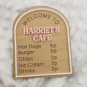 Puede incluir: Un letrero de madera arqueado con un borde blanco que presenta el texto WELCOME TO HARRIET'S CAFE en letras rosas en relieve. Debajo, un men&uacute; enumera Hot Dogs 5p, Burger 5p, Chips 3p, Ice Cream 3p y Drinks 2p en texto negro grabado.