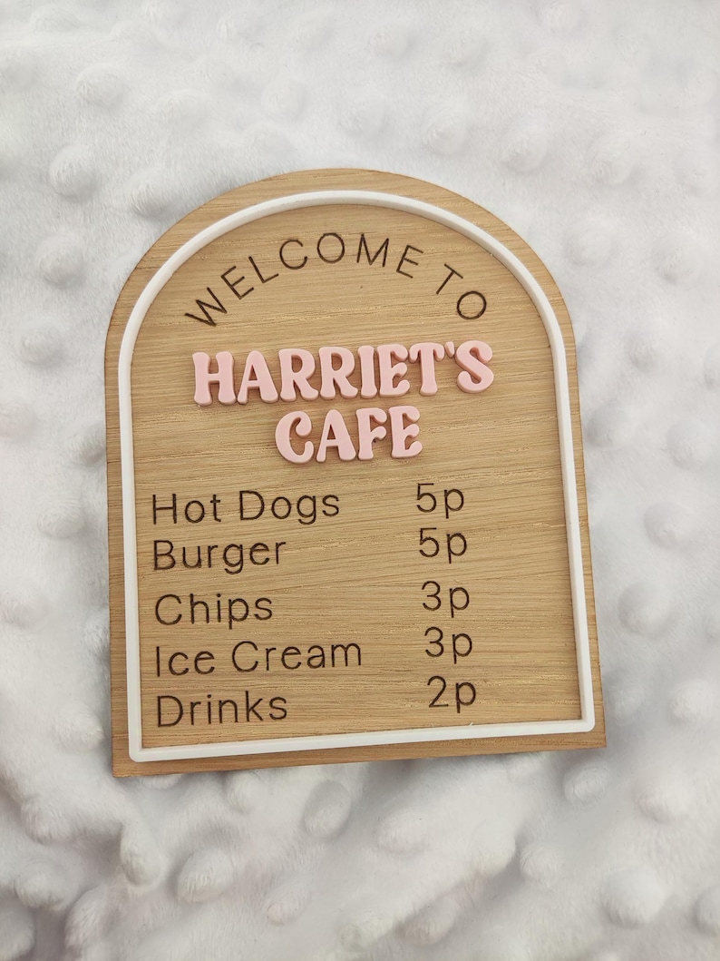 Puede incluir: Un letrero de madera arqueado con un borde blanco que presenta el texto WELCOME TO HARRIET'S CAFE en letras rosas en relieve. Debajo, un men&uacute; enumera Hot Dogs 5p, Burger 5p, Chips 3p, Ice Cream 3p y Drinks 2p en texto negro grabado.