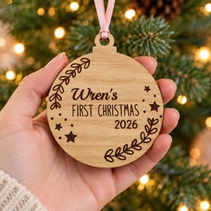 Puede incluir: Un adorno navideño redondo de madera con una cinta rosa claro. La superficie está grabada con el texto Wren's FIRST CHRISTMAS 2026, rodeado de ramas decorativas con hojas y pequeños iconos de estrellas en los lados izquierdo y derecho.