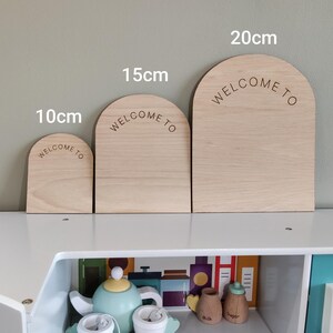 Puede incluir: Tres letreros de madera de color claro en forma de arco con el texto WELCOME TO grabado en cada uno. Los letreros est&aacute;n etiquetados con alturas de 10cm, 15cm y 20cm. Las piezas de madera tienen un acabado liso y un borde superior redondeado.