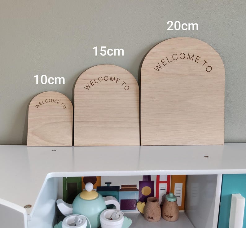 Puede incluir: Tres letreros de madera de color claro en forma de arco con el texto WELCOME TO grabado en cada uno. Los letreros est&aacute;n etiquetados con alturas de 10cm, 15cm y 20cm. Las piezas de madera tienen un acabado liso y un borde superior redondeado.