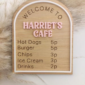 Puede incluir: Un letrero de madera arqueado con borde blanco que presenta el texto WELCOME TO HARRIET'S CAFE en letras rosas en relieve. Debajo, un men&uacute; enumera Hot Dogs 5p, Burger 5p, Chips 3p, Ice Cream 3p y Drinks 2p en letras negras grabadas.