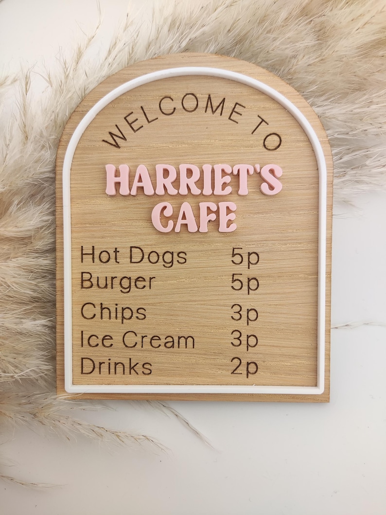 Puede incluir: Un letrero de madera arqueado con borde blanco que presenta el texto WELCOME TO HARRIET'S CAFE en letras rosas en relieve. Debajo, un men&uacute; enumera Hot Dogs 5p, Burger 5p, Chips 3p, Ice Cream 3p y Drinks 2p en letras negras grabadas.