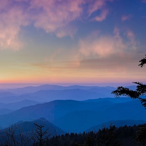 May include: A panoramic view of a mountain range at sunset. The sky is a vibrant mix of pink, purple, and orange, with wispy clouds. The sun is setting behind the mountains, casting a warm glow over the landscape. The mountains are layered in shades of blue and purple, with a hazy mist in the valleys. Silhouettes of trees stand tall in the foreground.