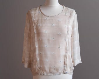May include: A vintage-style, cream-colored blouse with three-quarter sleeves and delicate floral embroidery. The sheer fabric drapes over a mannequin, showcasing the garment's details. The blouse has a round neckline and vertical paneling.