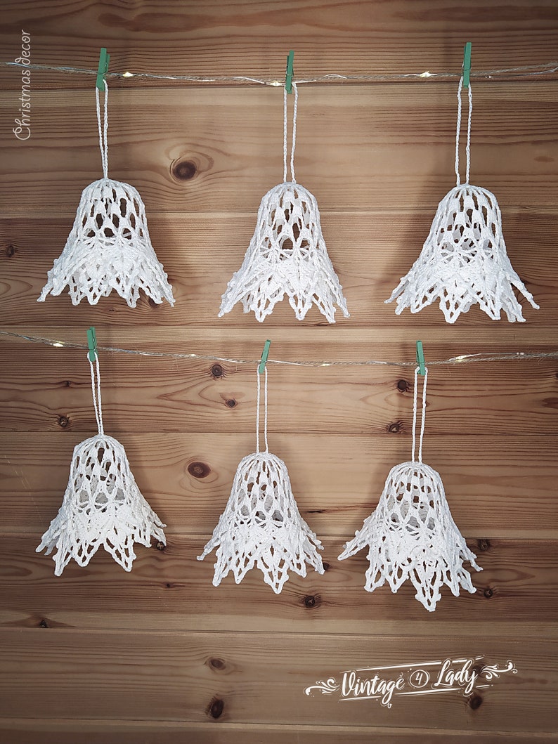 May include: Six white crocheted bells hanging from a string with green clothespins on a wood background. The bells are decorated with a lacy pattern. The text "Vintage Lady" is at the bottom of the image.