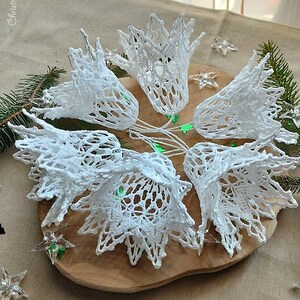 May include: A set of six white crocheted bells, arranged in a circle on a wooden surface. The bells are delicate and intricate, with a lacy design. The bells are perfect for adding a touch of vintage charm to your Christmas decor.