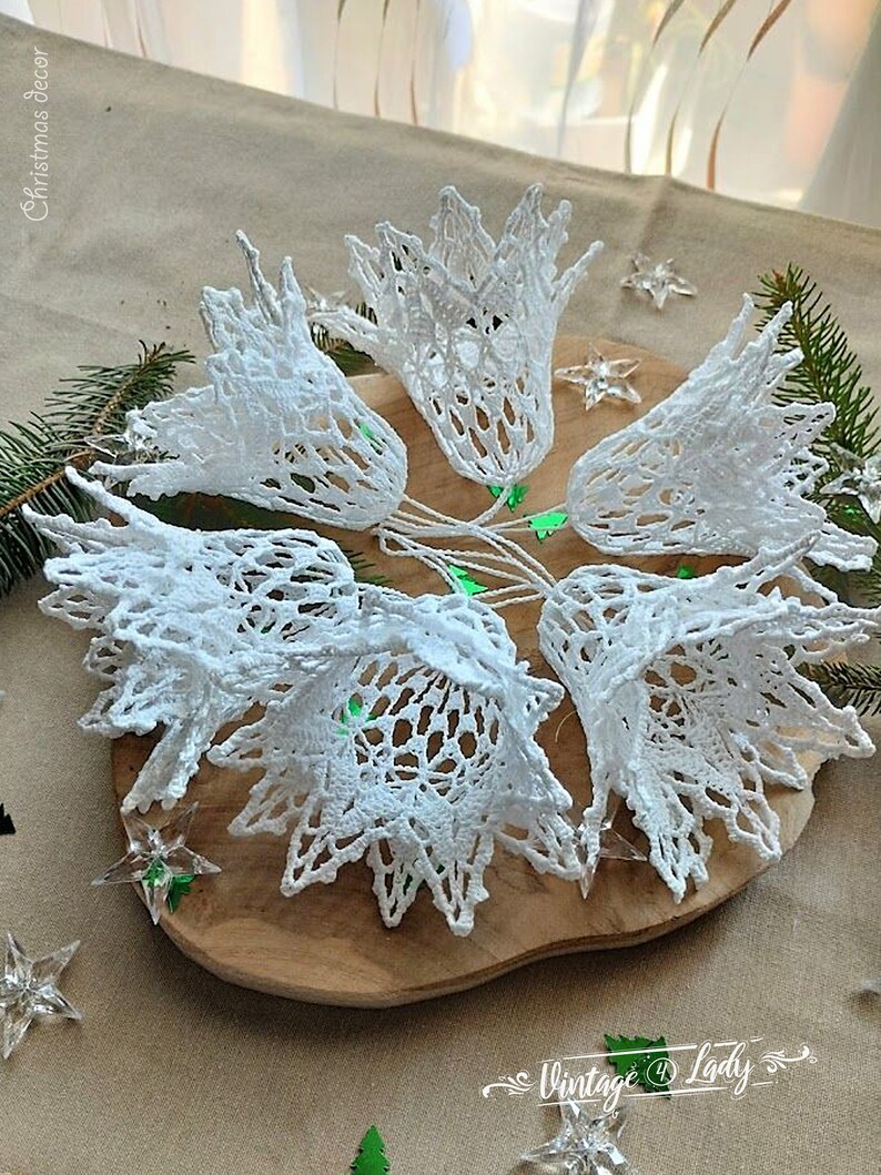 May include: A set of six white crocheted bells, arranged in a circle on a wooden surface. The bells are delicate and intricate, with a lacy design. The bells are perfect for adding a touch of vintage charm to your Christmas decor.