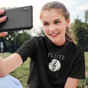 May include: A woman wearing a black t-shirt with a white graphic of a treble clef and the word "FLUTE" on it. She is taking a selfie with a black smartphone.