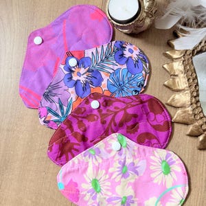 Peut inclure: Quatre serviettes hygiéniques lavables en tissu avec boutons-pression. La première est violet uni, la deuxième présente un motif floral bleu et orange, la troisième est magenta foncé avec un imprimé de feuilles, et la dernière a un motif de marguerites roses.