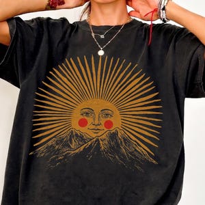 May include: Black t-shirt featuring a sun graphic with a face, radiating lines, and mountain imagery. The sun is in shades of gold and the face has red cheeks. The shirt has short sleeves and a relaxed fit.