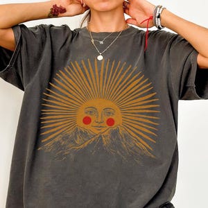 May include: Dark gray t-shirt featuring a sun graphic with a smiling face and mountain range. The sun has golden rays and red cheeks. The shirt has short sleeves and a relaxed fit.