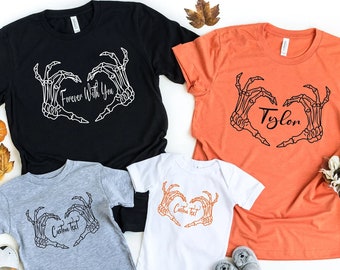 May include: A set of four t-shirts featuring a skeleton hand heart design. The shirts are in black, orange, gray, and white. The black shirt says "Forever With You", the orange shirt says "Tyler", and the gray and white shirts say "Custom Text".