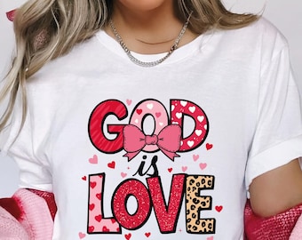 May include: White t-shirt with the text "GOD is LOVE" in red, pink, and leopard print. The word "GOD" has a heart pattern, and the word "LOVE" has a leopard print. A pink bow is between the words. Small red hearts are scattered around the text.