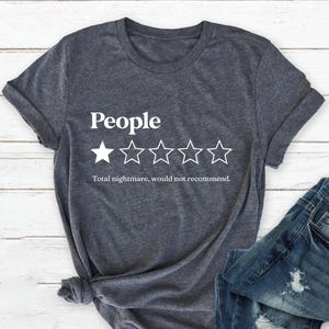 May include: Dark gray t-shirt with the word "People" above a one-star rating and the text "Total nightmare, would not recommend." The shirt is paired with blue denim jeans.