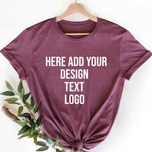 May include: A maroon short-sleeved t-shirt hanging on a wooden hanger. The center of the shirt features white text that reads: HERE ADD YOUR DESIGN TEXT LOGO. The fabric has a heathered texture, and a small green leafy branch is tucked underneath.