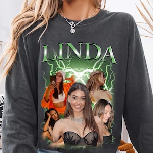 May include: Dark gray long-sleeve sweatshirt featuring the name "LINDA" in green with a lightning bolt design. The graphic includes multiple images of women. The sweatshirt is made of a soft material.