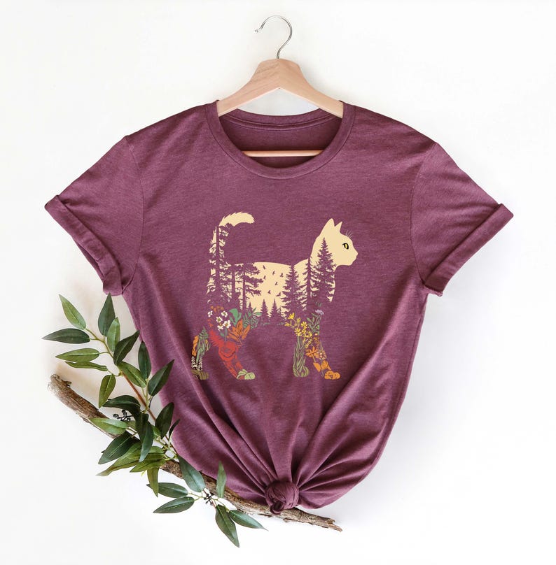 May include: A maroon t-shirt featuring a cat silhouette filled with a forest design. The cat's body is filled with trees, flowers, and foliage in various colors. The shirt is hanging on a wooden hanger.
