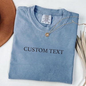 May include: A dusty blue short-sleeve t-shirt with a crew neckline and the black text CUSTOM TEXT printed on the chest. The shirt features a Comfort Colors brand label at the inner neck, and a gold-toned chain necklace rests on the fabric.