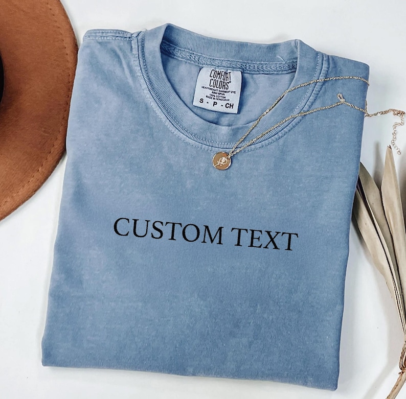 May include: A dusty blue short-sleeve t-shirt with a crew neckline and the black text CUSTOM TEXT printed on the chest. The shirt features a Comfort Colors brand label at the inner neck, and a gold-toned chain necklace rests on the fabric.