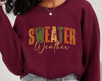 May include: A burgundy sweatshirt with the words "SWEATER WEATHER" in a distressed, multi-colored font. The word "Sweater" is in orange, green, and orange, while "Weather" is in a cursive, golden-yellow font.