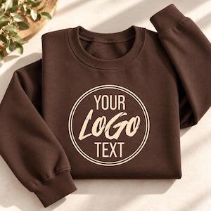 May include: A dark brown crewneck sweatshirt featuring a circular graphic on the chest. Inside the double-lined circle, the text reads YOUR LOGO TEXT in a combination of bold sans-serif and stylized brush-stroke lettering in a light cream color.