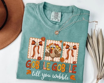 May include: A teal Comfort Colors t-shirt featuring a Thanksgiving-themed graphic. The design includes a turkey, bows, and the phrase "GOBBLE GOBBLE till you wobble" in colorful lettering. A silver necklace with a pendant is also visible.