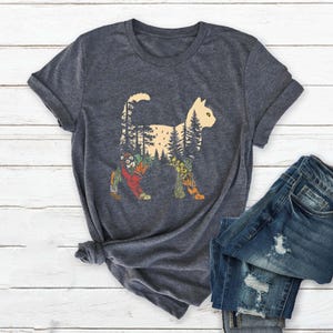 May include: Dark gray t-shirt featuring a cat silhouette filled with a forest scene. The cat design includes trees, flowers, and other natural elements in shades of green, orange, and yellow. The shirt is paired with blue denim jeans.