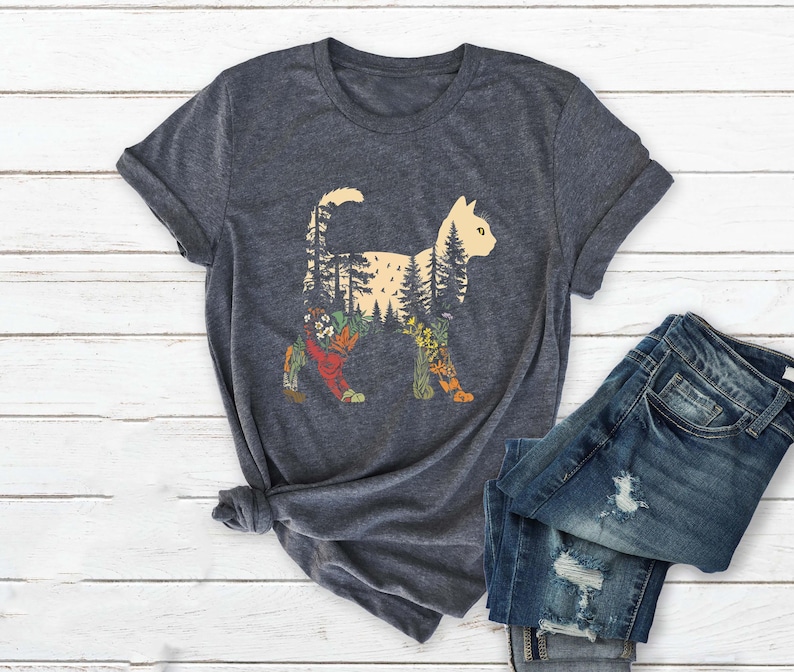May include: Dark gray t-shirt featuring a cat silhouette filled with a forest scene. The cat design includes trees, flowers, and other natural elements in shades of green, orange, and yellow. The shirt is paired with blue denim jeans.