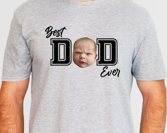 May include: A light gray t-shirt with the words "Best Dad Ever" in black script and block letters. The letter "D" features a baby's face. The shirt is short-sleeved.
