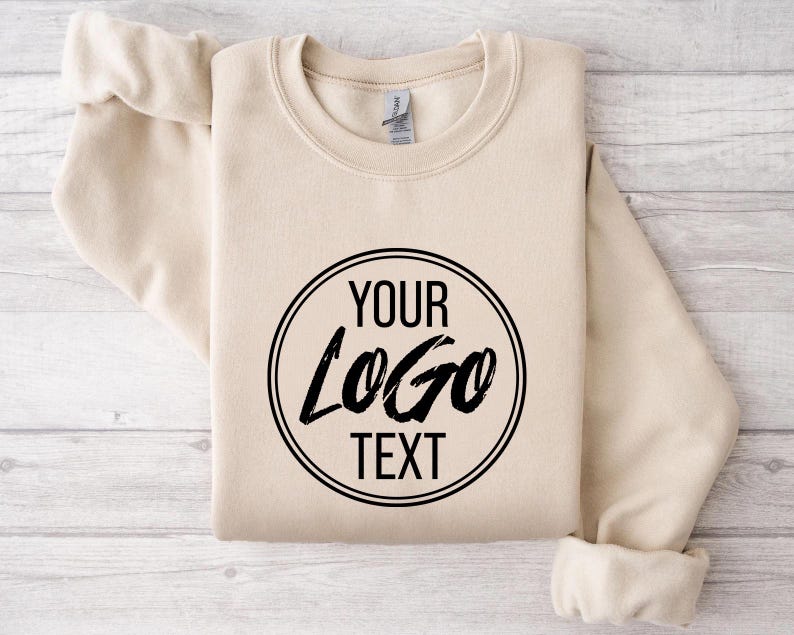 May include: A sand-colored crewneck sweatshirt featuring a black circular graphic on the chest. Inside the circle, the text reads YOUR LOGO TEXT in a combination of bold sans-serif and stylized brush-stroke lettering. The garment has long sleeves.