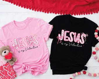 May include: Two t-shirts, one pink and one black, with the text "JESUS is my Valentine" in a patchwork design. The word "JESUS" is in pink and white patterns. Pink bows and stars accent the text. A teddy bear and Valentine's Day decorations are in the background.