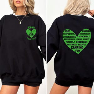 May include: Black sweatshirt with a white collar. The front features a green heart-shaped graphic with the name "SOPHIA", a tent, and "TROOP 25077". The back displays a green heart with words like "Camping Fun", "Kindness", and "Courageous".