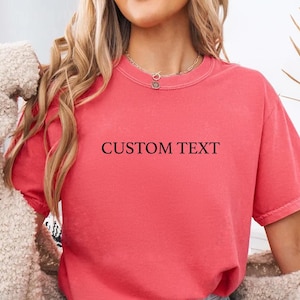 May include: A coral-colored short-sleeve t-shirt featuring the words CUSTOM TEXT printed in a simple black serif font across the center of the chest. The garment has a crew neckline and a relaxed fit, shown with a beige textured cardigan.