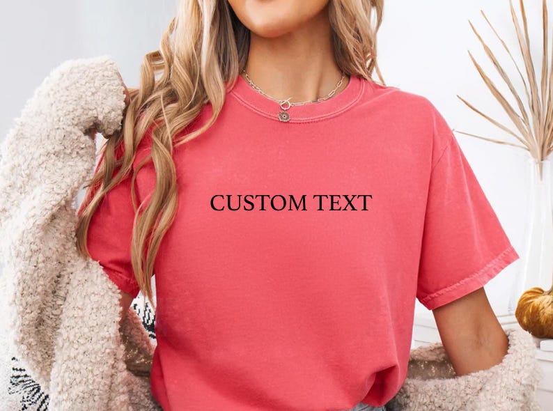 May include: A coral-colored short-sleeve t-shirt featuring the words CUSTOM TEXT printed in a simple black serif font across the center of the chest. The garment has a crew neckline and a relaxed fit, shown with a beige textured cardigan.