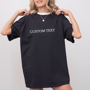 May include: A black short-sleeved t-shirt featuring the words CUSTOM TEXT printed in white capital letters across the chest. The garment has a crew neckline and a loose, relaxed fit, providing a simple and straightforward design for casual wear.