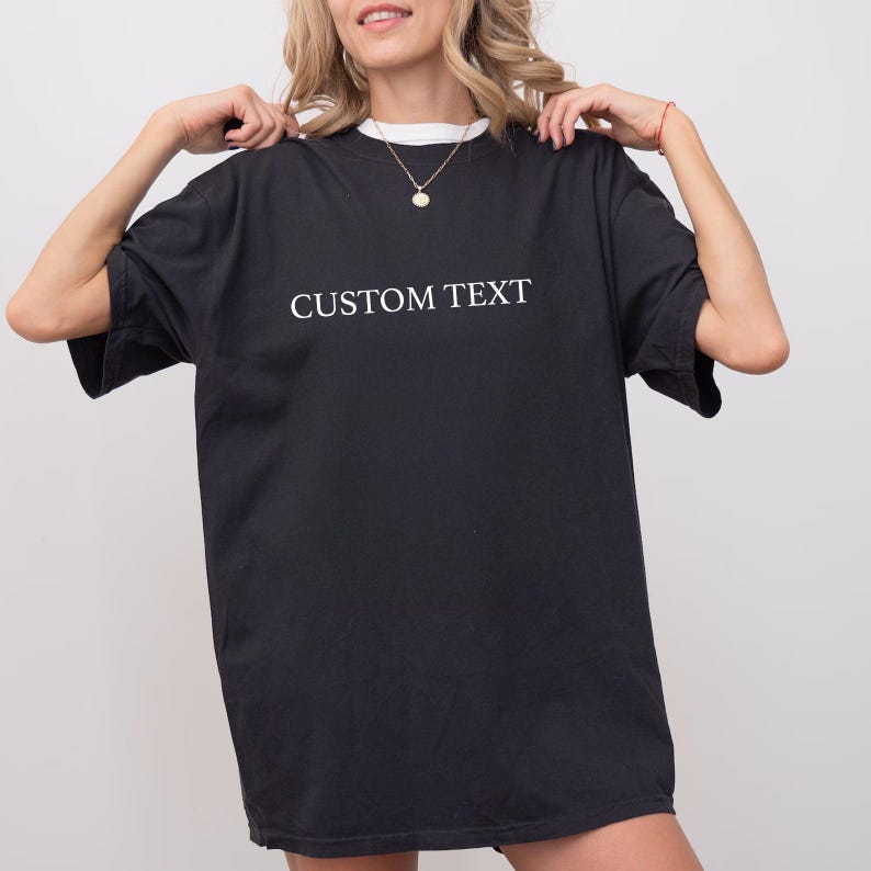 May include: A black short-sleeved t-shirt featuring the words CUSTOM TEXT printed in white capital letters across the chest. The garment has a crew neckline and a loose, relaxed fit, providing a simple and straightforward design for casual wear.