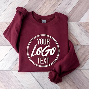 May include: A maroon long-sleeve crewneck sweatshirt featuring a white circular graphic on the chest. Inside the circle, the text reads YOUR LOGO TEXT in a bold, stylized font. The garment has a ribbed collar, cuffs, and hem, with a Gildan brand label.