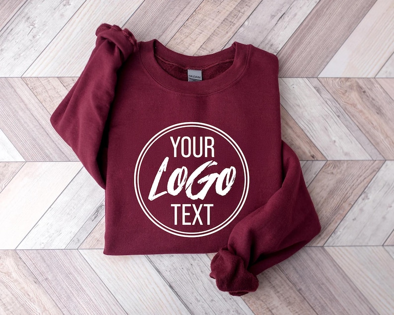 May include: A maroon long-sleeve crewneck sweatshirt featuring a white circular graphic on the chest. Inside the circle, the text reads YOUR LOGO TEXT in a bold, stylized font. The garment has a ribbed collar, cuffs, and hem, with a Gildan brand label.