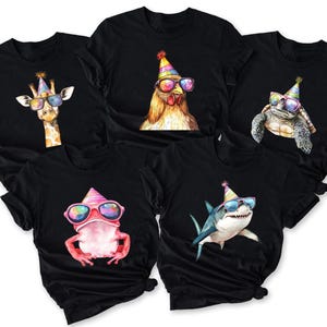 May include: Five black short-sleeve t-shirts, each featuring a different animal wearing a colorful party hat and rainbow-tinted sunglasses. The animals include a giraffe, a chicken, a turtle, a pink frog, and a shark, all rendered in a vibrant, artistic style.