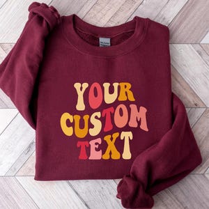 May include: Burgundy sweatshirt with the text "YOUR CUSTOM TEXT" in a retro font with orange, yellow, red, and pink colors.