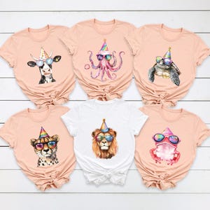 May include: Five peach-colored t-shirts and one white t-shirt, each featuring a watercolor illustration of an animal wearing a colorful party hat and sunglasses. The animals include a cow, an octopus, a turtle, a cheetah, a lion, and a frog.