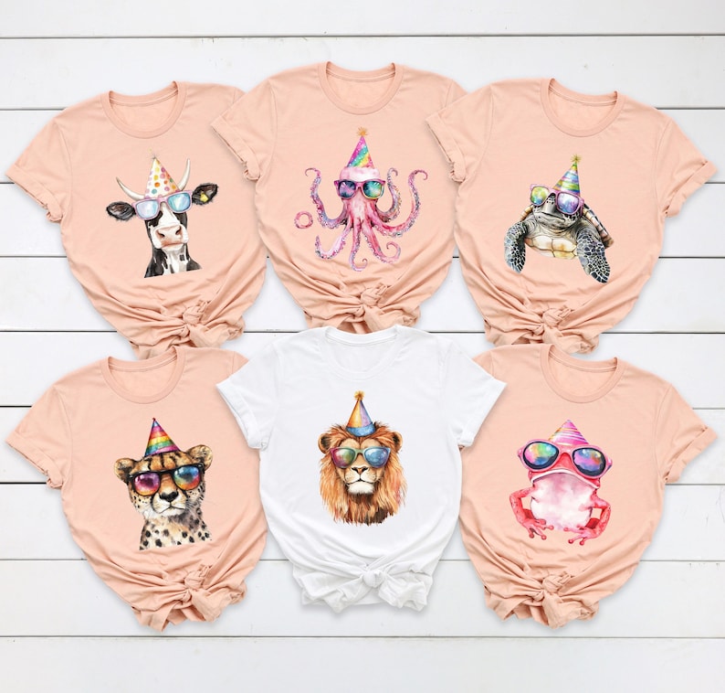 May include: Five peach-colored t-shirts and one white t-shirt, each featuring a watercolor illustration of an animal wearing a colorful party hat and sunglasses. The animals include a cow, an octopus, a turtle, a cheetah, a lion, and a frog.