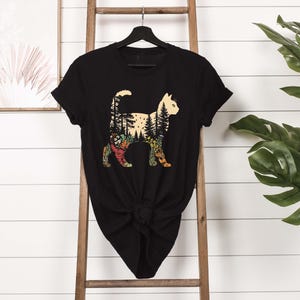 May include: Black t-shirt featuring a cat silhouette with a forest design inside. The cat's body is filled with trees, flowers, and foliage in shades of green, yellow, and orange. The shirt is hanging on a black hanger.