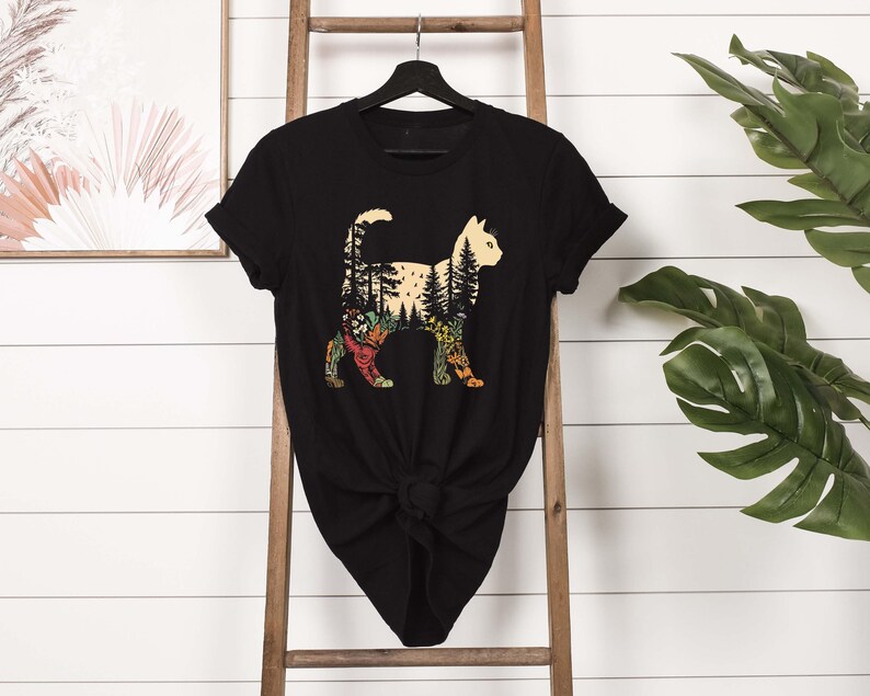 May include: Black t-shirt featuring a cat silhouette with a forest design inside. The cat's body is filled with trees, flowers, and foliage in shades of green, yellow, and orange. The shirt is hanging on a black hanger.