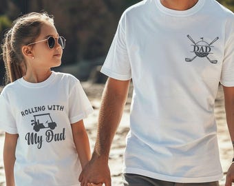 May include: White t-shirts with golf-themed graphics. One shirt says "Rolling with My Dad" with a golf cart illustration. The other shirt has a golf ball, clubs, and the word "DAD".