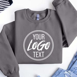 May include: A charcoal gray crewneck sweatshirt featuring a white circular graphic on the chest. Inside the circle, the text reads YOUR LOGO TEXT in a combination of sans-serif and brush-style fonts. The garment has long sleeves and a ribbed collar.