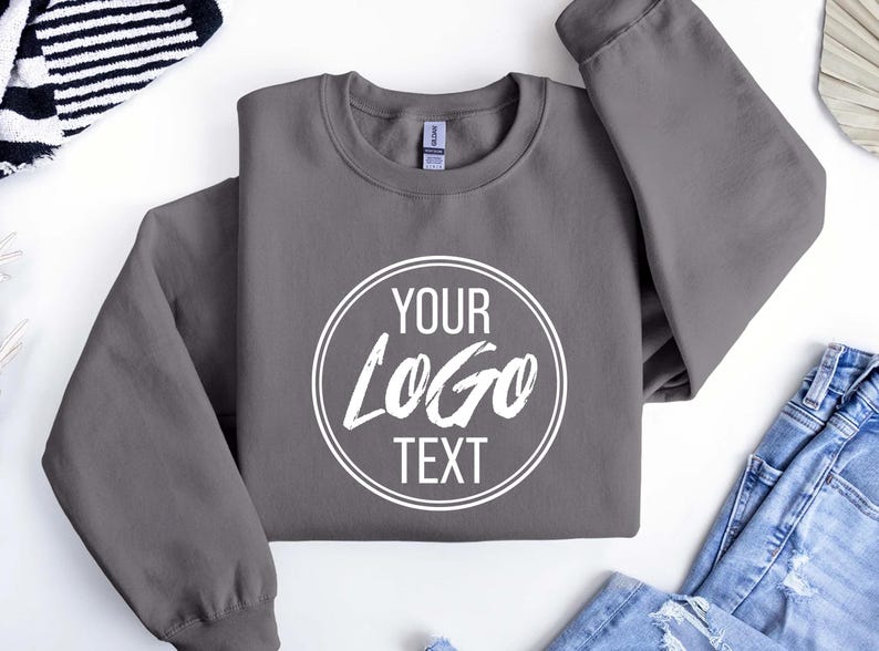 May include: A charcoal gray crewneck sweatshirt featuring a white circular graphic on the chest. Inside the circle, the text reads YOUR LOGO TEXT in a combination of sans-serif and brush-style fonts. The garment has long sleeves and a ribbed collar.