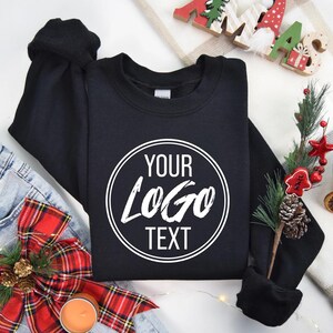 May include: A black crewneck sweatshirt featuring a white circular graphic on the chest with the text "YOUR LOGO TEXT" printed in a bold, sans-serif font. The sweatshirt has long sleeves and a ribbed collar, cuffs, and hem.